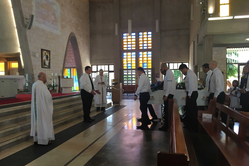 Men in white shirts carry a small white coffin to the front of a church.