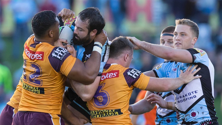 A rugby league player holds a football as he stands held up by an opposition player in a tackle.