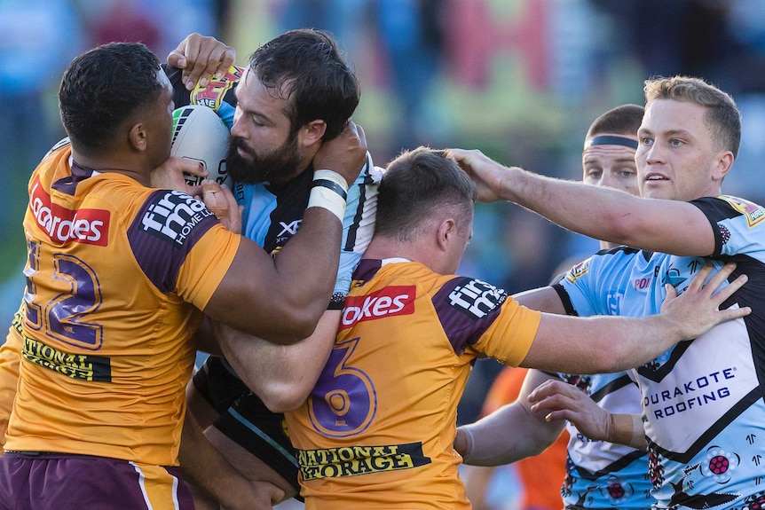 A rugby league player holds a football as he stands held up by an opposition player in a tackle.