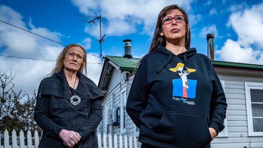Two women stand outside a house, looking serious.