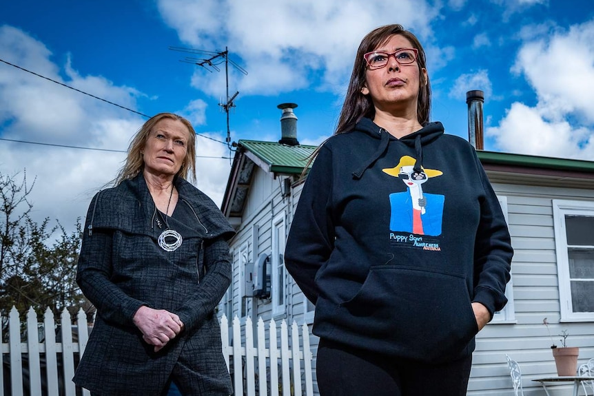 Two women stand outside a house, looking serious.