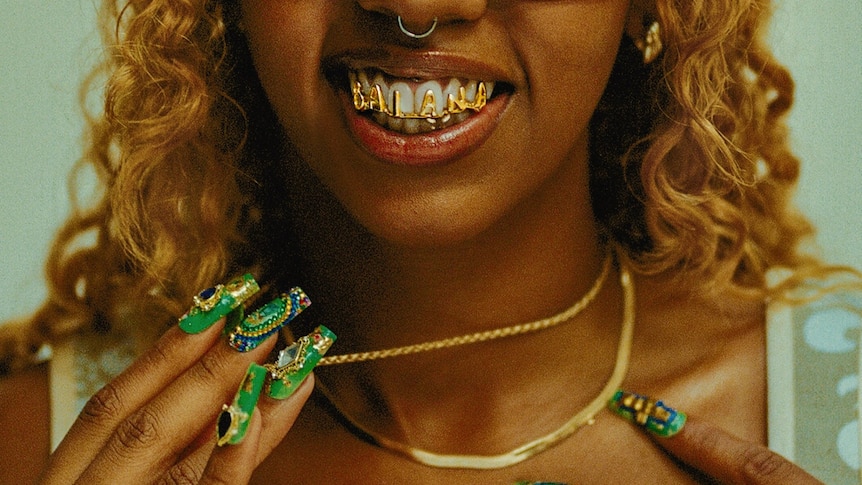 A woman with green jewelled nails is baring her teeth. She has a gold grill with the word "Baianá. You cannot see her eyes.