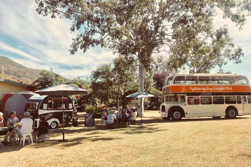 People sit under umbrellas and trees with a double decker bus parked nearby