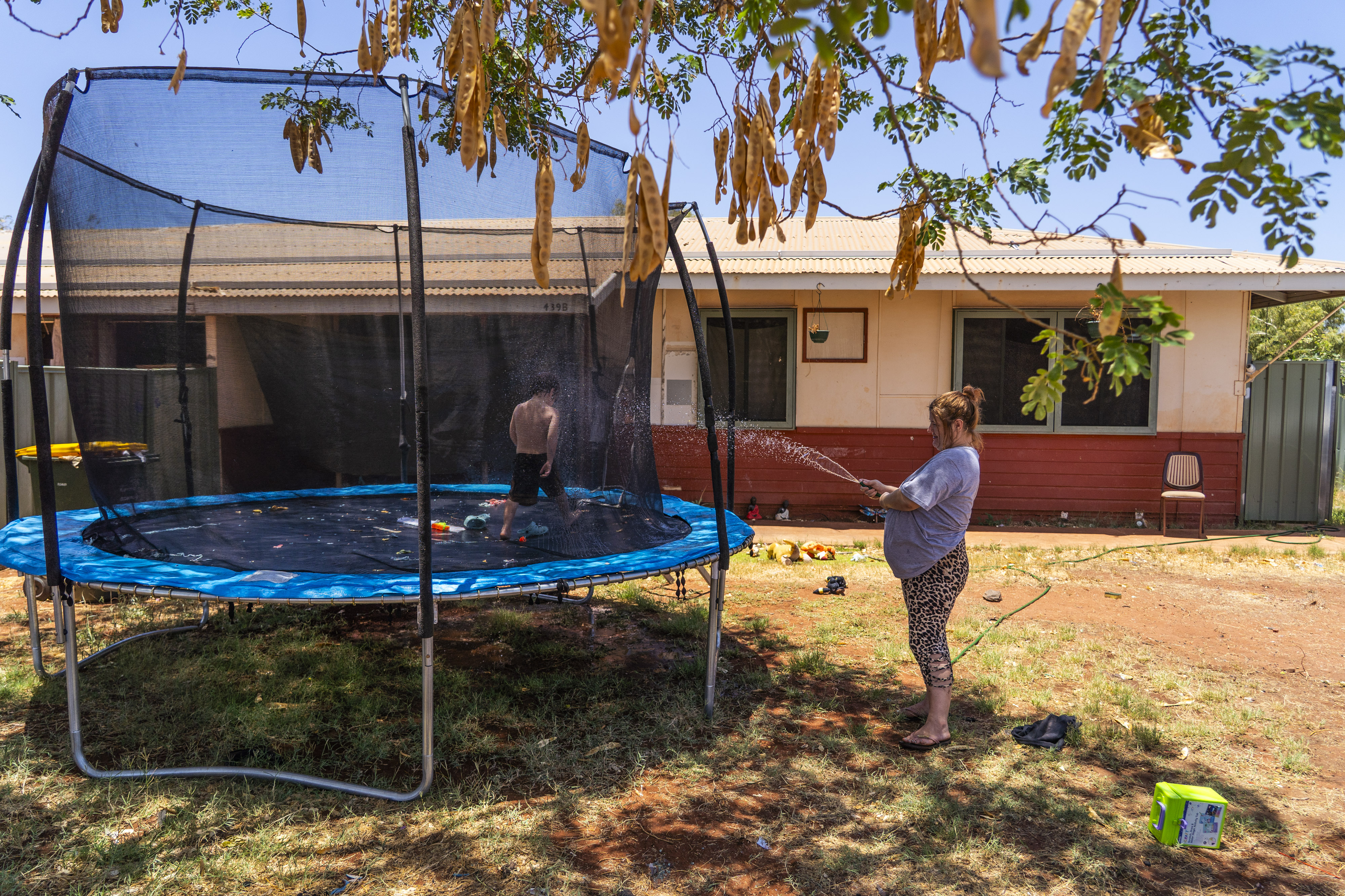 Uma criança brinca num trampolim à sombra enquanto a mãe borrifa água.