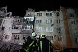 Two firefighters examine a large apartment block damaged by shelling, with destroyed windows and cratered upper storeys.