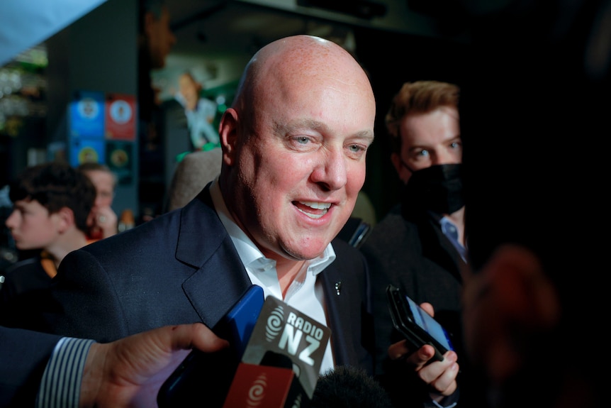 A bald man stands in front of several news microphones