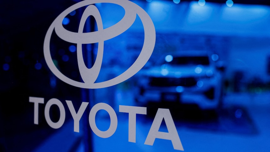 A Toyota logo sits on a blue glass window display in grey writing with a car behind it