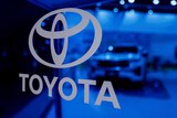 A Toyota logo sits on a blue glass window display in grey writing with a car behind it
