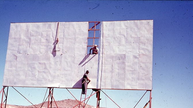 Three men construct a large white screen, with one on a ladder looking back at the camera. Red desert surround the screen