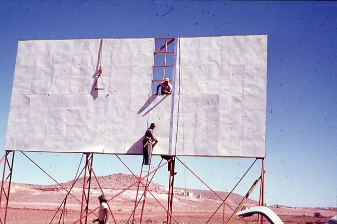 Three men construct a large white screen, with one on a ladder looking back at the camera. Red desert surround the screen