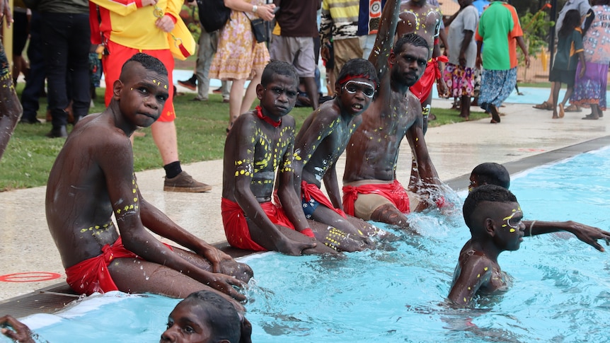 Kids in traditional attire and face paint sit with their feet in a swimming pool. Children splash around them.