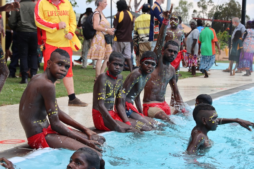 Kids in traditional attire and face paint sit with their feet in a swimming pool. Children splash around them.