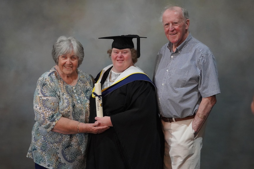 A woman with Down syndrome wearing an academic gown and holding a rolled up certificate with her elderly parents 