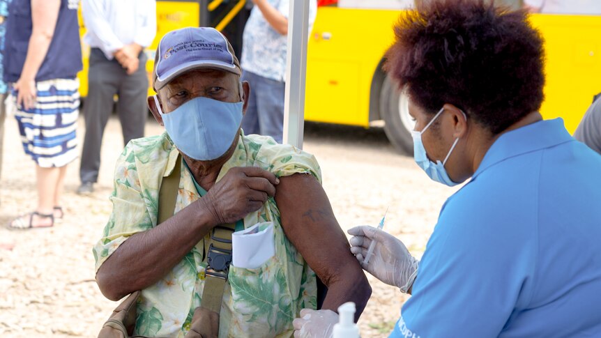 A Papua New Guinean man in a blue face mask holds up a sleeve so a nurse can inject his shoulder