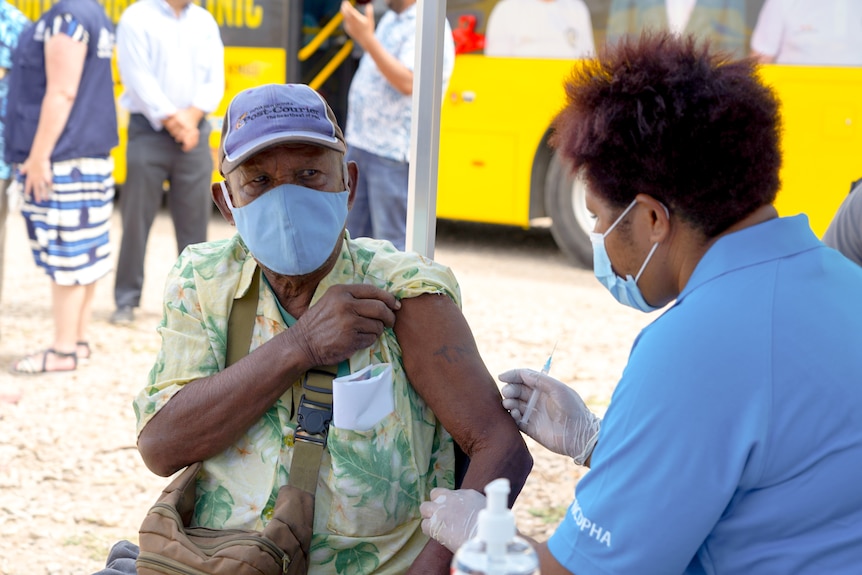 A Papua New Guinean man in a blue face mask holds up a sleeve so a nurse can inject his shoulder