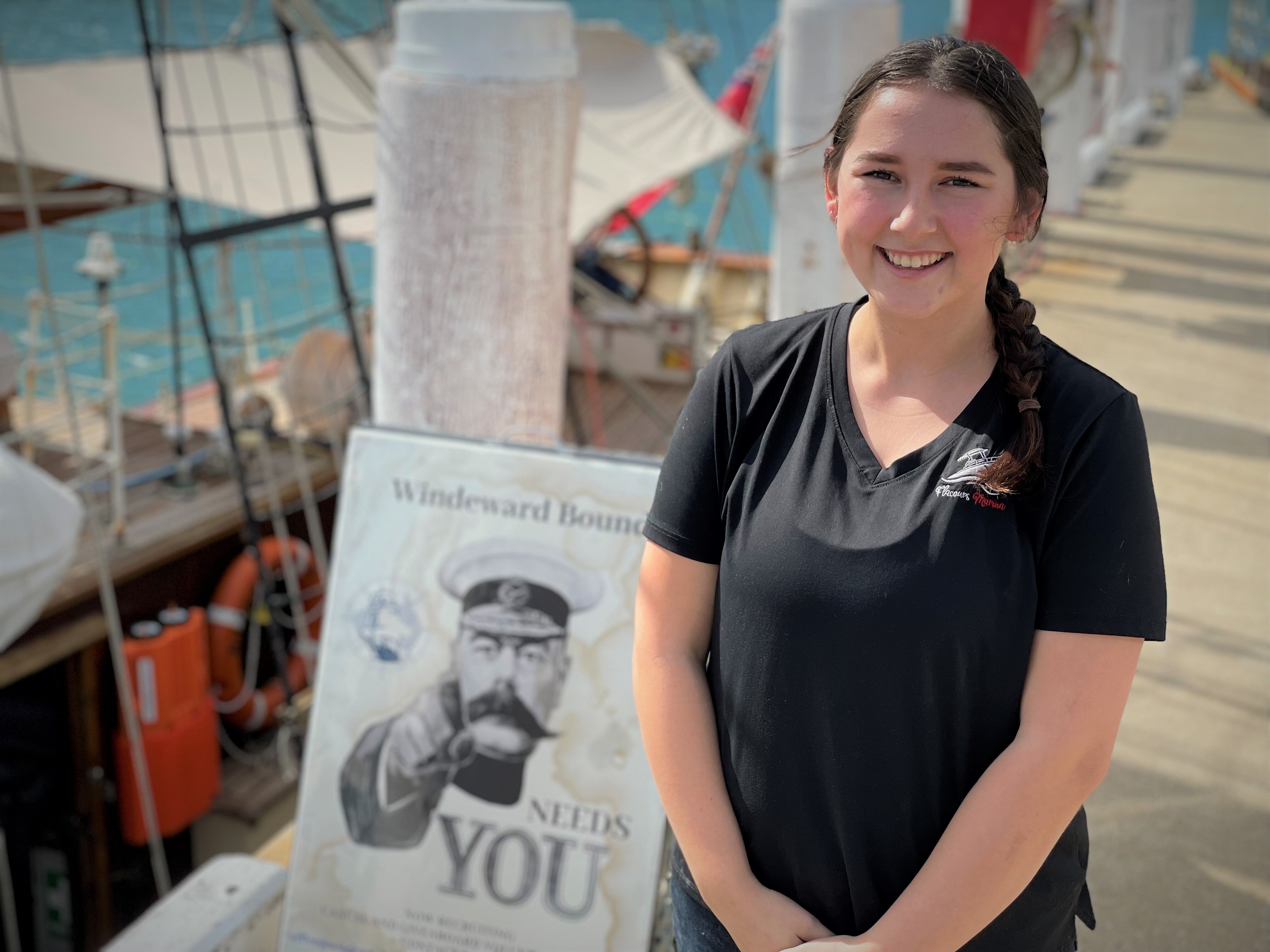 A brunette girl in a black T-shirt smiling at the camera standing on a jetty next to a ship and sign