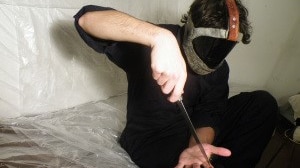 A masked man wearing black clothes sitting down on the floor cuts one of his fingers with a knife.