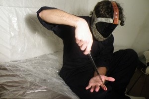 A masked man wearing black clothes sitting down on the floor cuts one of his fingers with a knife.