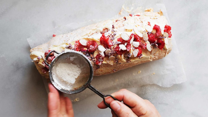 Dusting icing sugar over a pavlova roll topped with cream, raspberries and almonds for a festive dessert recipe.