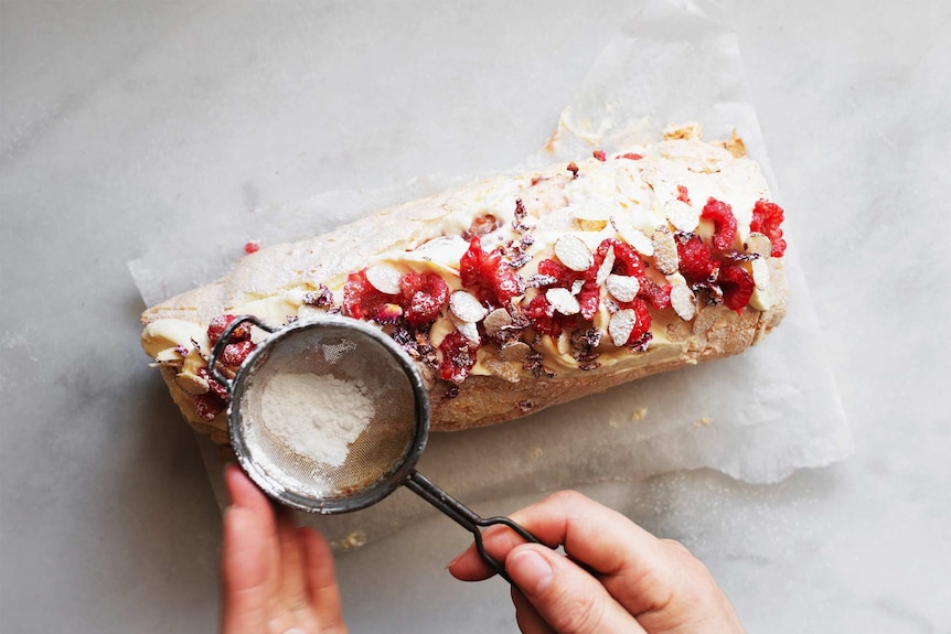 Dusting icing sugar over a pavlova roll topped with cream, raspberries and almonds for a festive dessert recipe.