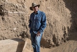 A man in double denim and fedora stands in an archaeological site. 