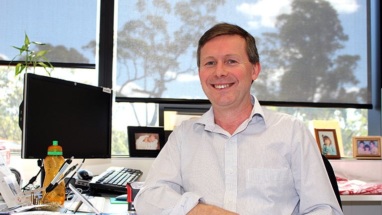 A man in a white shirt sits at a desk, with a computer and family photographs behind him.