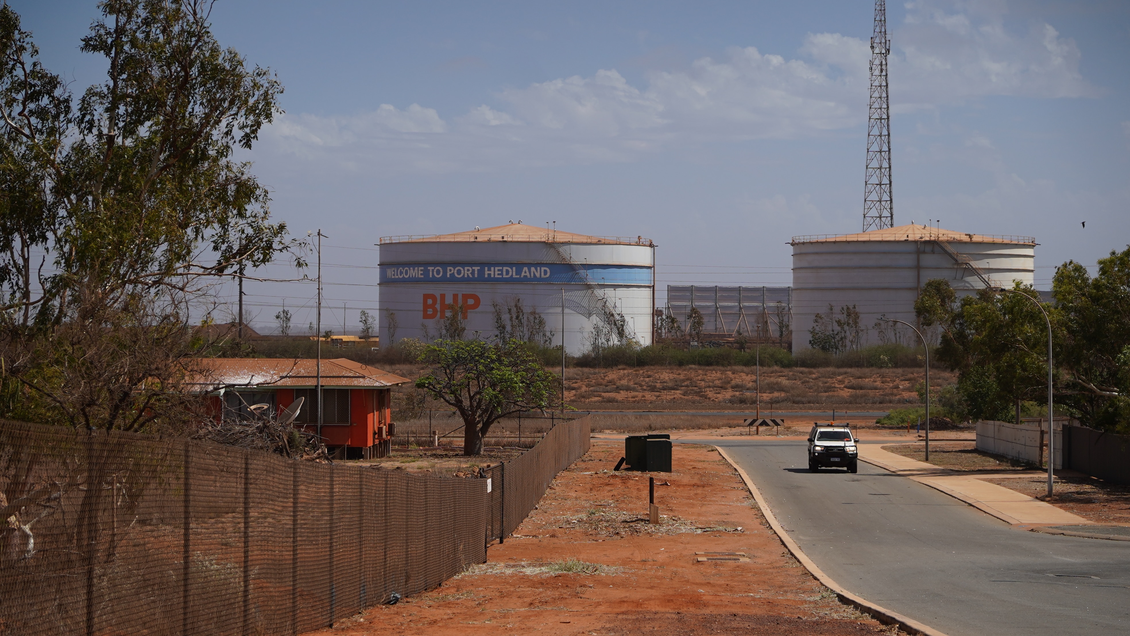 Una carretera con un automóvil conduciendo junto con un gran tanque de agua de BHP al fondo y polvo naranja a lo largo de la franja natural.
