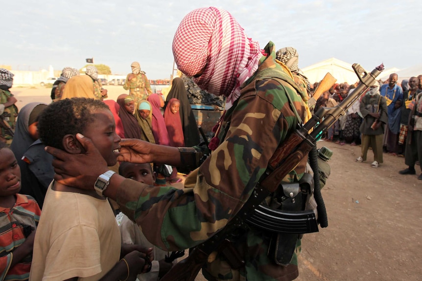 A man in army fatigues with his face obscured by a scarf gently puts his hands on a boy's face