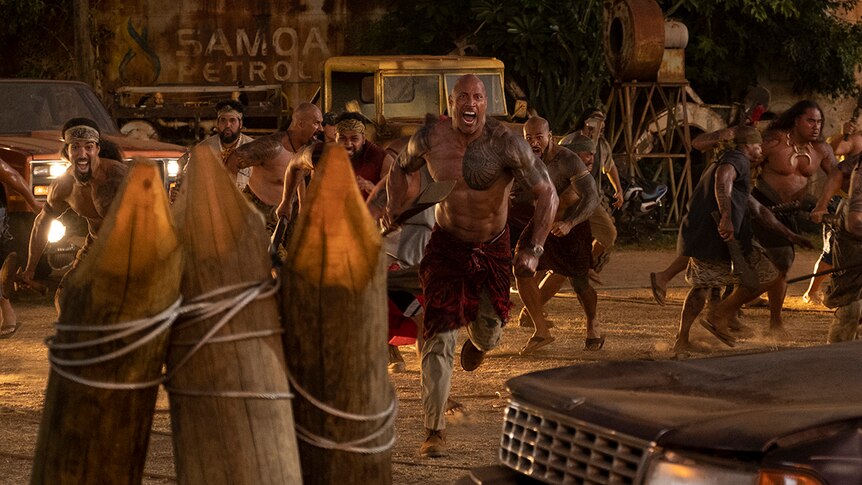 A small group of muscular men with traditional Polynesian tattoos charge forwards with weapons in industrial area near cars.