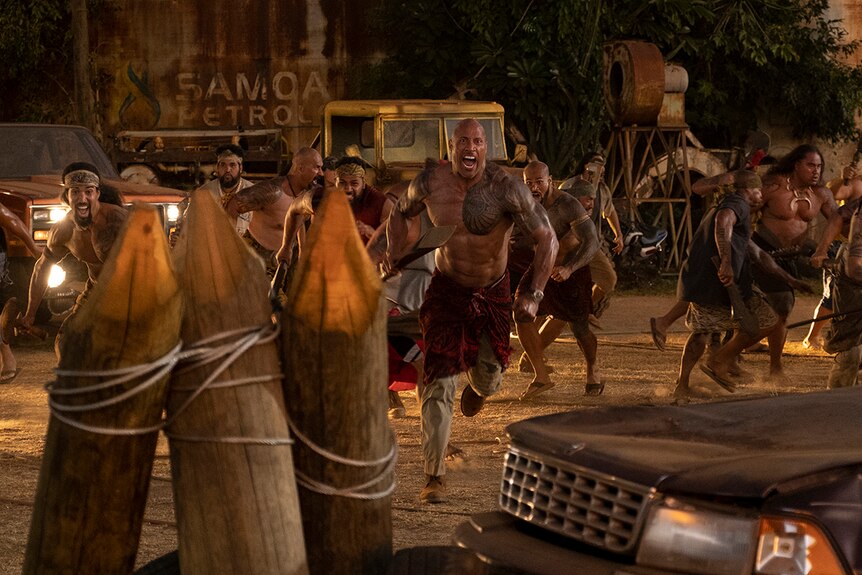 A small group of muscular men with traditional Polynesian tattoos charge forwards with weapons in industrial area near cars.