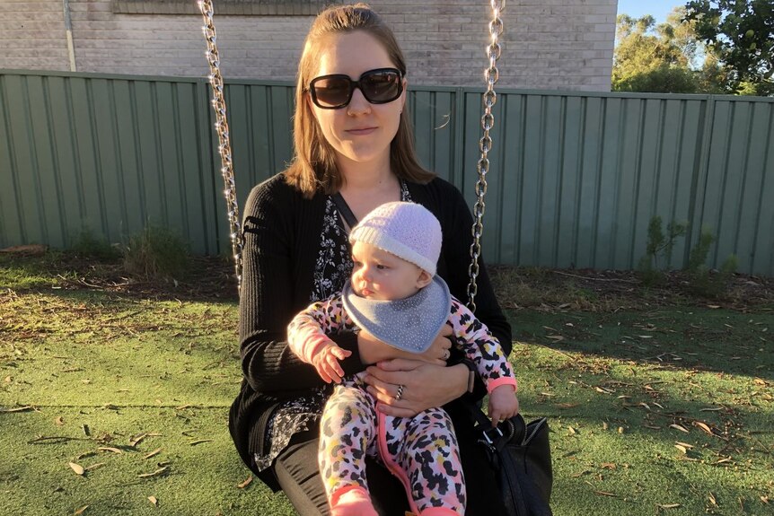 A woman in sunglasses sitting on a swing holding a baby in a beanie.