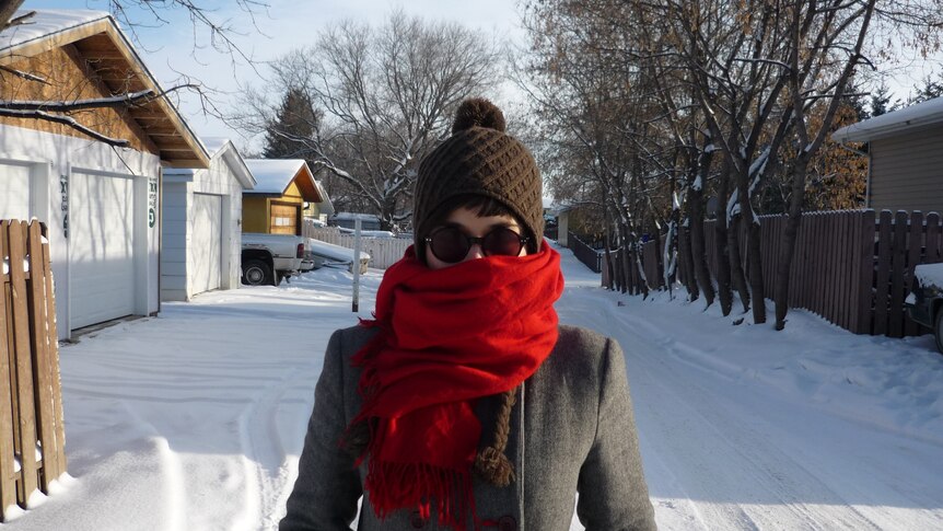 A woman on a snow covered road wearing a brown beanie, red scarf, glasses and a gray jacket
