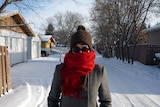 A woman on a snow covered road wearing a brown beanie, red scarf, glasses and a gray jacket