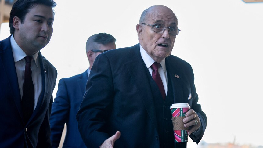 An elderly man standing and walkin in a suit holding a cup of coffee and with a surprised look on his face. Two men behind.