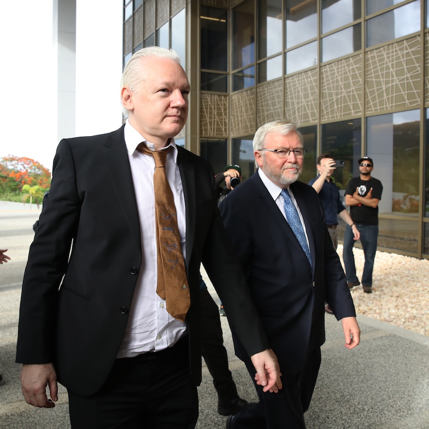 Two middle-aged white men in suits walks past reporters outside a building.