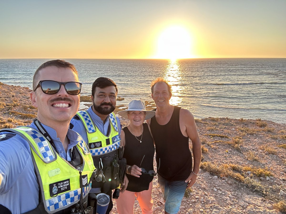 Two police officers take a selfie photo with two people next to the ocean at sunset.