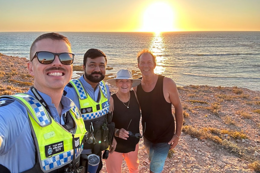Two police officers take a selfie photo with two people next to the ocean at sunset.