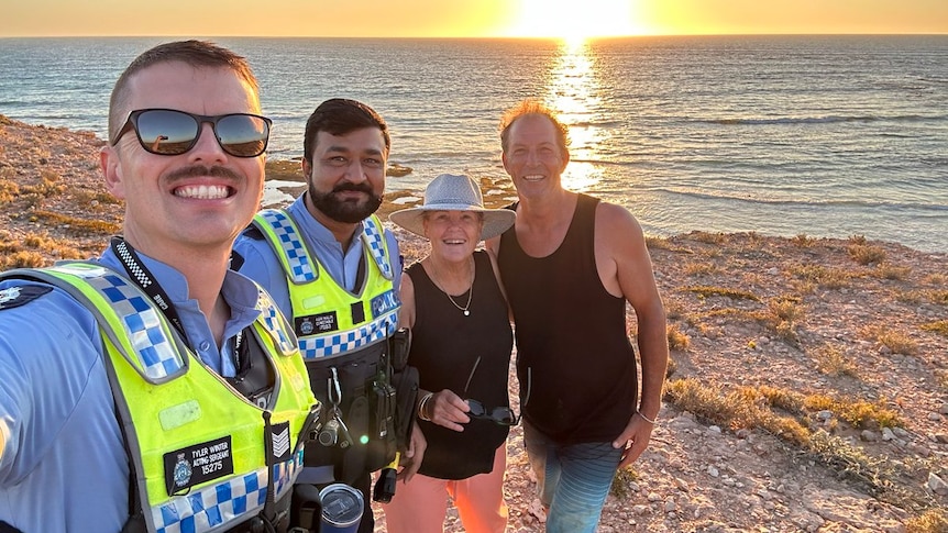 Two police officers take a selfie photo with a smiling man and woman next to the ocean at sunset.
