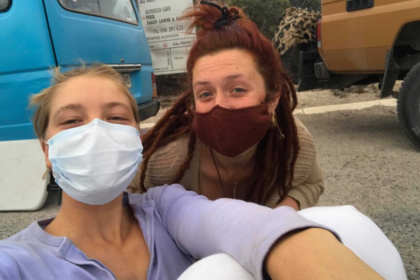 Two women wearing masks in a selfie crouched down in front of vehicles.