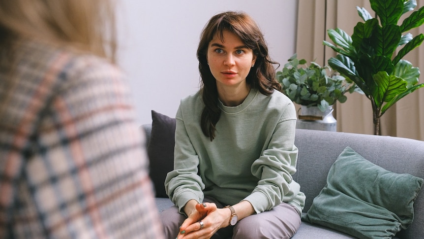 A young woman sits on a couch with her hands clasped, speaking to a blurred taller woman in a tweed blazer.