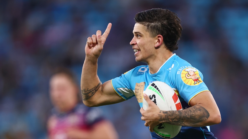 A Gold Coast Titans player grins and points his finger to the sky as he runs back with the ball after scoring a try.