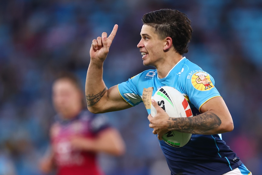 A Gold Coast Titans player grins and points his finger to the sky as he runs back with the ball after scoring a try.