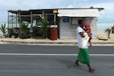 A mother carries a baby in front of a dilapidated shack