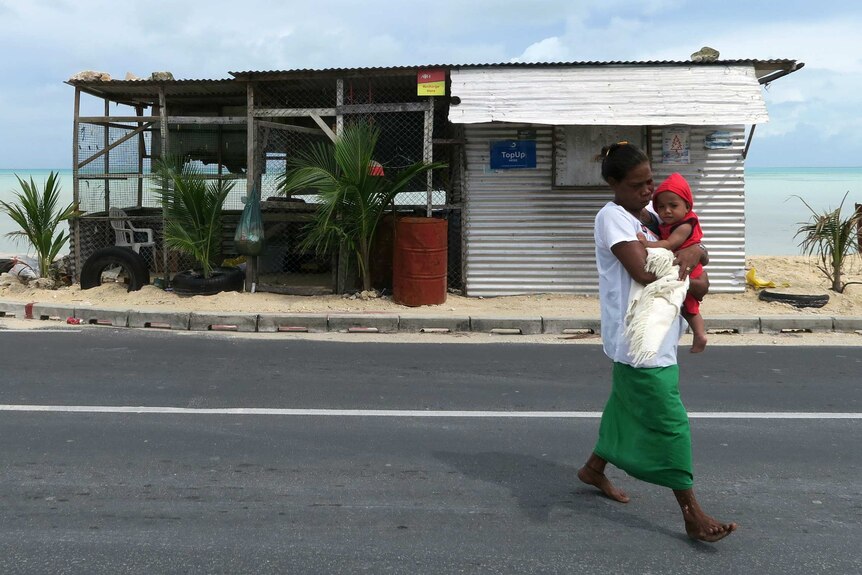 A mother carries a baby in front of a dilapidated shack