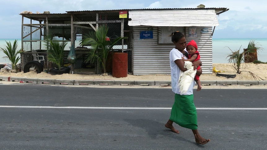 A mother carries a baby in front of a dilapidated shack