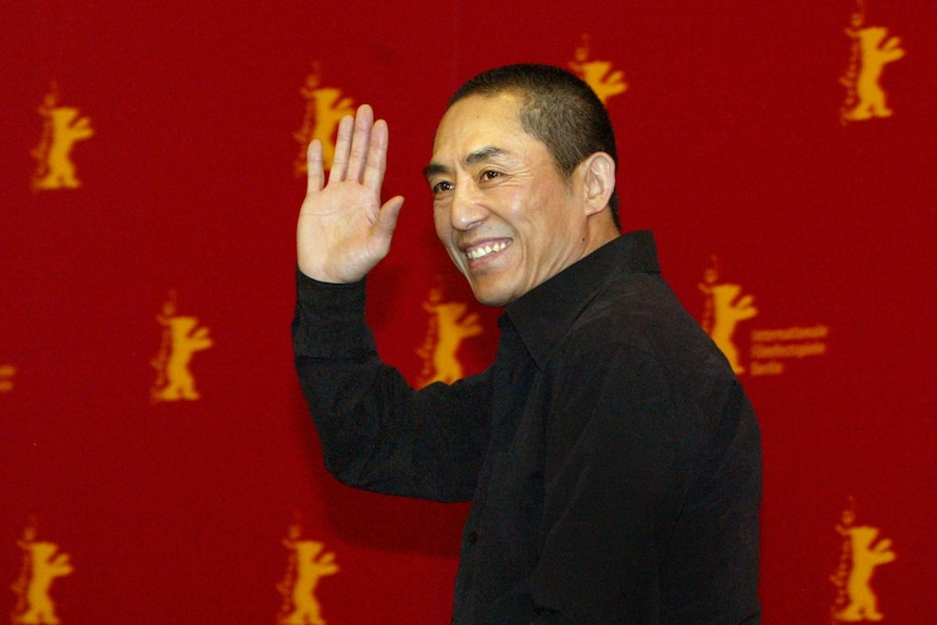 The director looks at the camera smiling and waving as he walks past a red backdrop illustrated with small yellow bears.