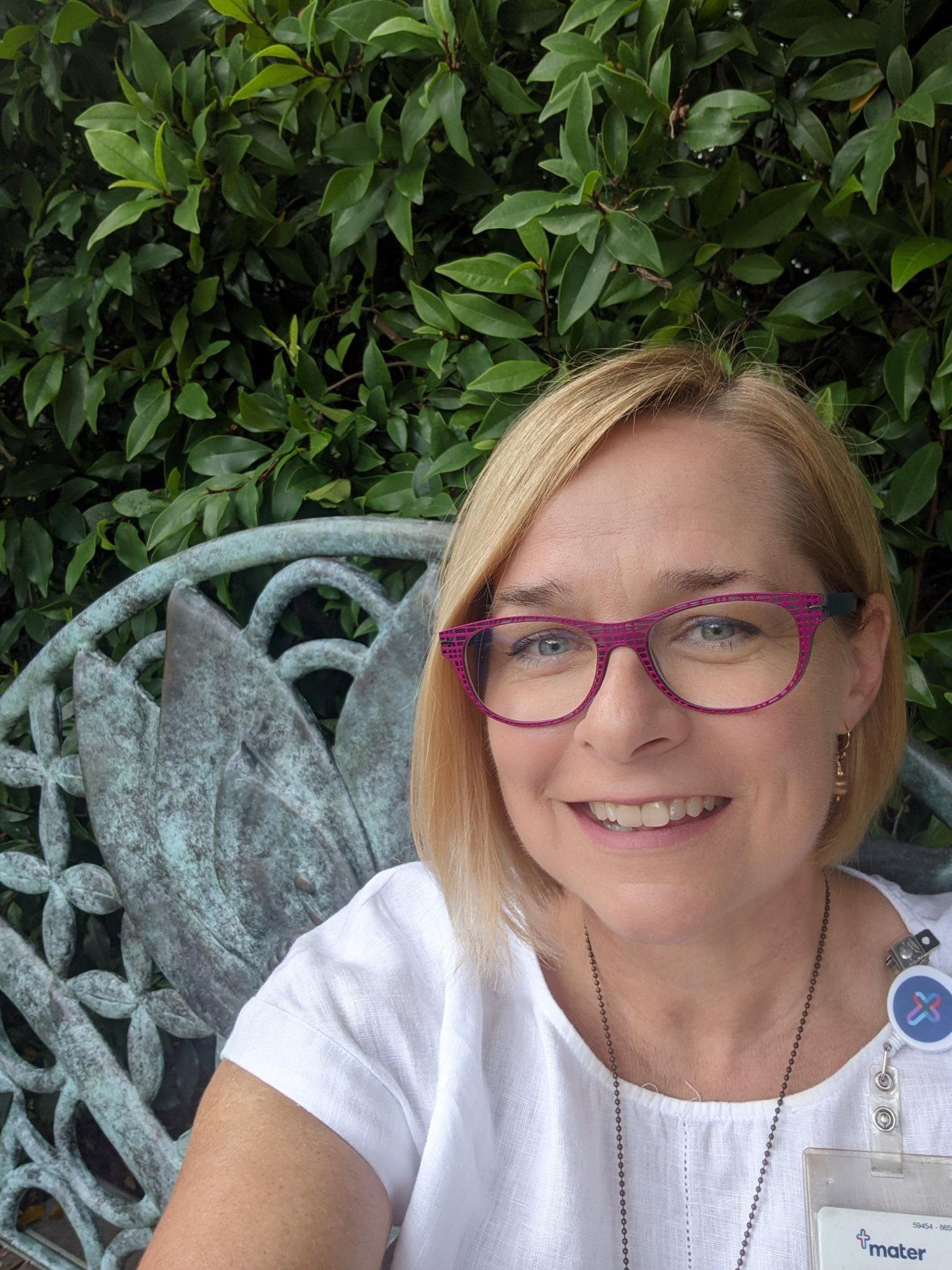 Woman wearing white t-shirt and red-framed glasses smiles at camera while sitting outside.