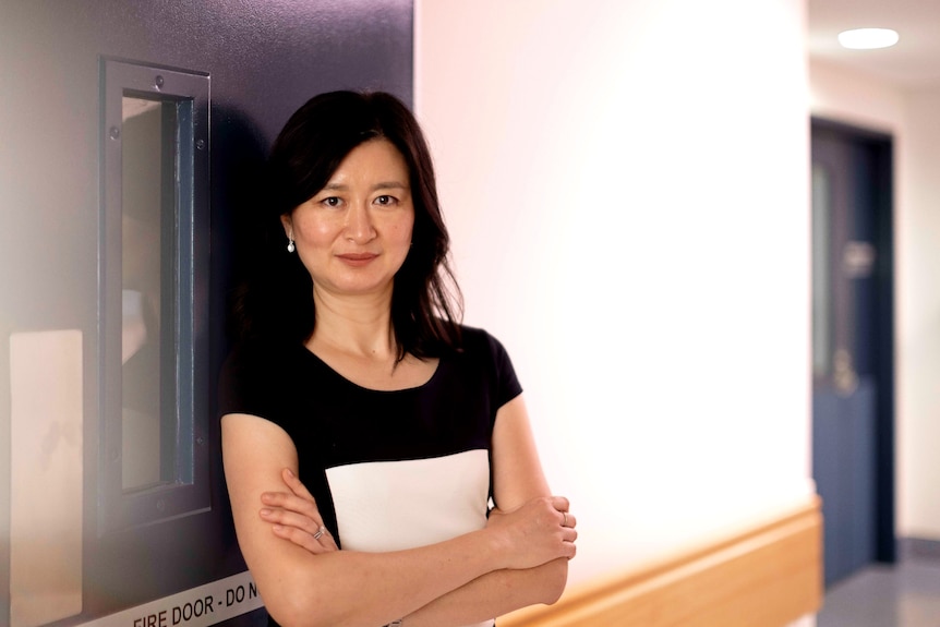 Christine Lin stands with her arms folded. She is leaning against a blue door in a corridor with white walls.