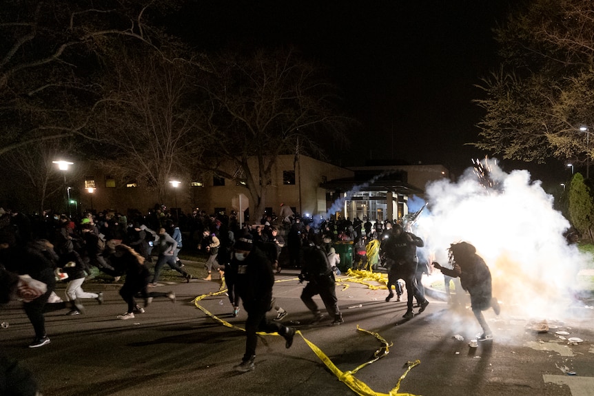 People flee police over downed cordon lines as smoke or gas billows from behind them.