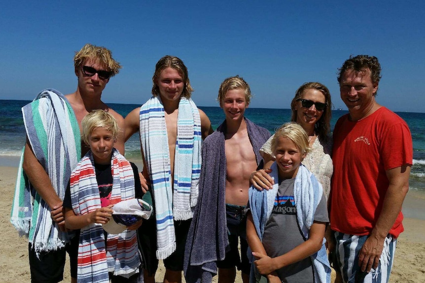 A family including mum and dad and five boys of various ages stands posing for a photo with towels on a beach.